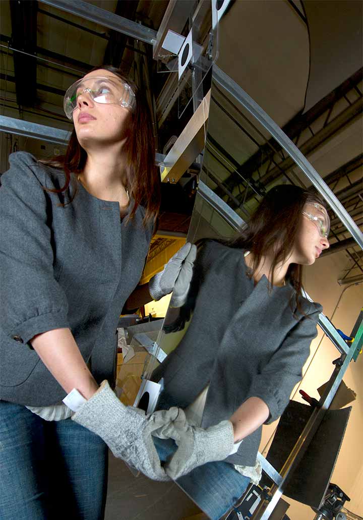 201909-facilities-29190 A woman in safety glasses is reflected in the large mirror she is moving with gloves.