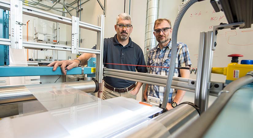 Two NRELians stand with roll-to-roll equipment in a laboratory.