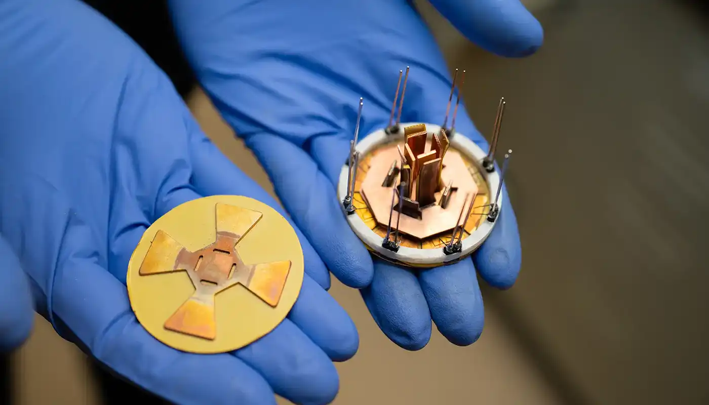 Two gloved hands hold a small octagonal power module and a corresponding attachment layer.